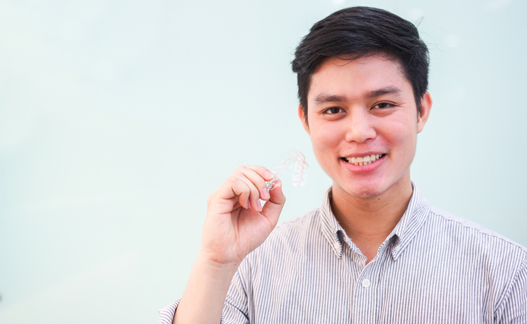 close up young asian man smiling with hand holding dental aligner retainer (invisible) at dental clinic for beautiful teeth treatment course concept - Dental News.