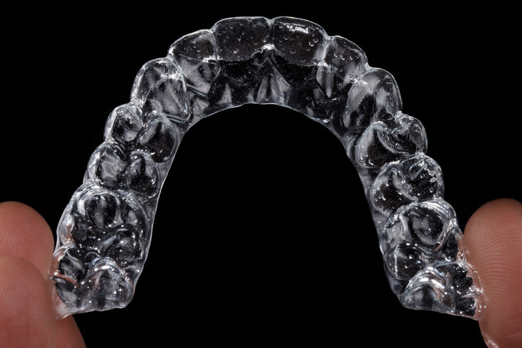 A close up of clear aligners held between two fingers in black background.