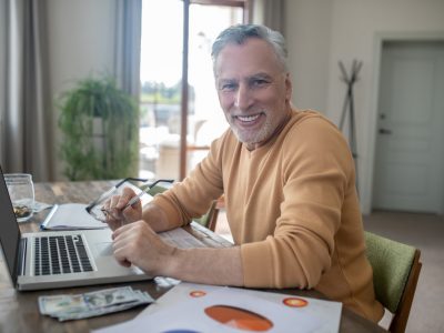 A grey-haired gentleman sitting at his desk with his laptop, smiling at the camera.