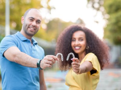 A man and woman standing beside one another, smiling, holding out their veneers for the viewer to see.