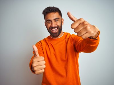 Young man wearing orange sweater over isolated white background approving doing positive gesture with hand, thumbs up smiling and happy for success. Winner gesture.
